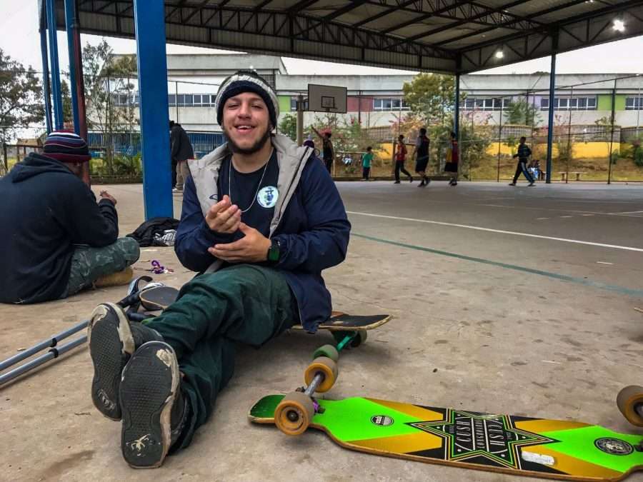 Marcos Vinícius Nascimento, que desenvolveu uma técnica especial para manter o equilíbrio em cima do skate usando órtese. Foto: Acervo pessoal Descrição: Homem jovem vestindo calça jeans, tênis, blusa de moletom e touca de lã está sentado em cima do skate. Do seu lado esquerdo estão suas muletas e, do lado direito, outro skate. Ao fundo, crianças brincam em uma quadra poliesportiva.