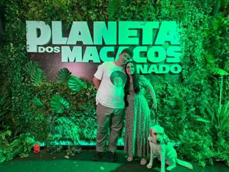 A foto mostra duas pessoas e um cachorro em frente a um painel com a inscrição "Planeta dos Macacos: Renascido". O painel está decorado com muitas plantas verdes, criando um ambiente de floresta. A pessoa à esquerda é um homem usando uma camiseta branca com uma estampa no centro e calças bege. Ele está com o braço em volta da mulher ao seu lado. A mulher está à direita, usando um vestido verde com bolinhas pretas e segurando a guia de um cachorro. O cachorro é um labrador amarelo, que está sentado no chão. A iluminação da foto é predominantemente verde, o que dá um tom esverdeado a toda a cena.