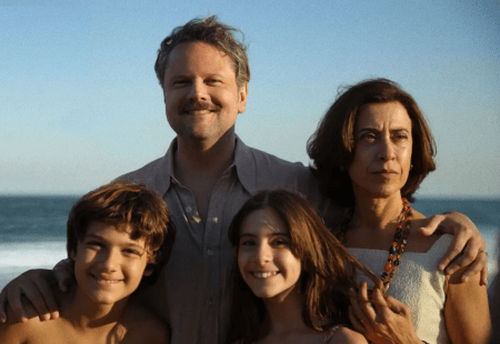 A imagem mostra uma família composta por quatro pessoas posando em frente ao mar. À esquerda, há um menino sorridente com cabelo castanho. Ao lado dele, uma menina também sorridente com cabelo castanho longo. Atrás das crianças, está um homem de meia-idade com cabelo castanho claro e bigode, sorrindo. À direita, uma mulher de meia-idade com cabelo castanho escuro, expressão séria, usando um colar colorido. O cenário é uma praia, com o mar ao fundo e o céu azul claro.
