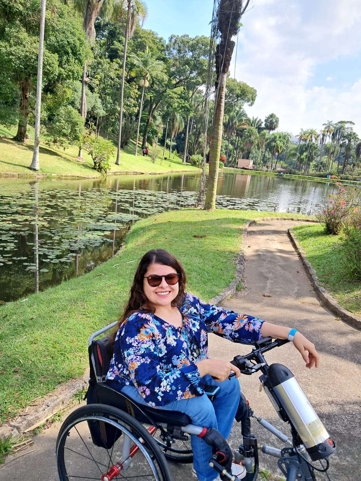 Mulher sorridente em cadeira de rodas motorizada, usando óculos escuros, blusa florida azul e calça jeans. Ela está em um parque arborizado, ao lado de um lago com muitas folhas de vitória-régia na superfície. O caminho é de cimento, rodeado por grama verde e árvores altas, incluindo palmeiras. O clima parece agradável, com céu parcialmente nublado e luz do sol suave. Ao fundo, há mais vegetação e um ambiente tranquilo e natural.