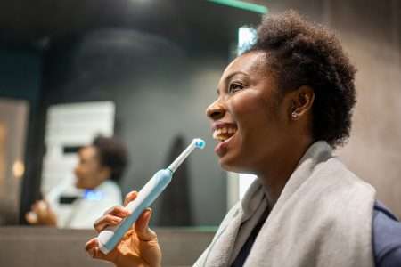 Mulher sorrindo enquanto escova os dentes com uma escova de dentes elétrica. Ela está em frente a um espelho no banheiro, usando um roupão ou toalha nos ombros. O ambiente é bem iluminado e moderno. A expressão da mulher é alegre e descontraída. No fundo, ela também aparece refletida no espelho.