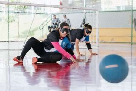 Fotografia: quadra de esportes indoor com duas mulheres usando vendas nos olhos, ajoelhadas perto de um gol. Elas estão jogando goalball, um esporte adaptado para pessoas com deficiência visual. À frente delas, há uma bola azul específica desse esporte, que possui guizos para que os jogadores possam ouvi-la. Ambas estão concentradas, com as mãos no chão, prontas para defender o gol. Ao fundo, uma rede protege a área do gol e há algumas pessoas sentadas, acompanhando a partida.