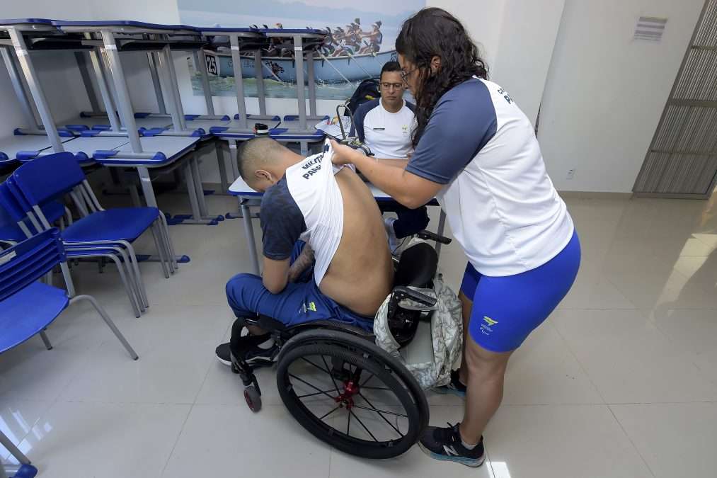 Fotografia: a imagem mostra uma cena de atendimento fisioterapêutico em que um homem sentado em uma cadeira de rodas está com a camiseta levantada enquanto uma mulher faz um procedimento nas costas dele, usando um aparelho portátil. Eles estão em uma sala com várias cadeiras azuis empilhadas e mesas. Outro homem, também com uniforme esportivo, observa a cena sentado ao fundo. Trata-se de um atendimento dirigido a atletas paralímpicos.