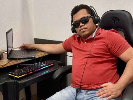 Um homem sentado numa cadeira gamer preta, em um canto de parede branca, ao lado de uma mesa de computador. Ele usa camiseta polo vermelha e jeans claro, está com fones de ouvido grandes e óculos escuros. Ele está reclinado, com a mão esquerda esticada apoiada perto do notebook aberto na mesa e a mão direita sobre a coxa. Na mesa há um notebook, um teclado com iluminação colorida (RGB) e cabos visíveis. A expressão e a postura passam um ar relaxado, como se estivesse descansando enquanto usa o computador.