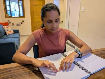 Uma mulher sentada à mesa lendo com as mãos um caderno em braille. Ela está em um ambiente interno, com expressão concentrada, olhando para baixo. O cabelo está preso para trás e ela usa uma blusa sem mangas, de gola alta, em tom rosado. No pulso direito há um relógio e uma pulseira. Sobre a mesa de madeira há um caderno grande de espiral aberto, com páginas brancas cheias de pontinhos em relevo (braille). As duas mãos estão apoiadas na página, com os dedos percorrendo as linhas. Ao fundo, à esquerda, aparece um sofá azul com almofadas coloridas; atrás dele, uma janela com grades e uma fileira de fotos pequenas penduradas. No centro há uma porta de madeira; à direita, uma parede clara e parte de um corredor/entrada.