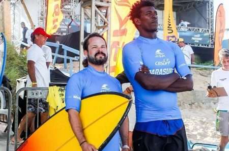 Dois surfistas vestindo camisetas azuis, ambos com a logo “CBSurf” e a palavra “Parasurf”. Eles estão juntos em uma praia, um deles segura uma prancha de surfe amarela com detalhes pretos, enquanto o outro está com os braços cruzados. Ao fundo, há outras pessoas, algumas bandeiras, uma estrutura metálica e areia, indicando o ambiente de competição. O clima parece ser descontraído e amistoso.