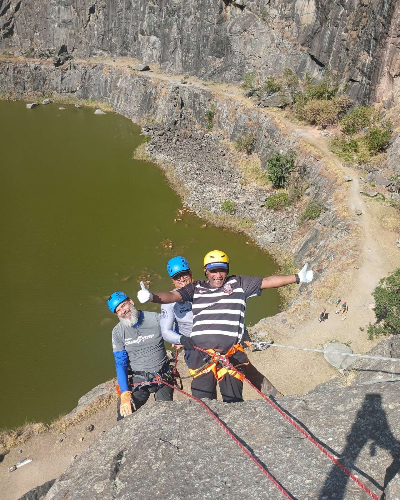 Três homens praticando rapel em uma rocha alta, com um lago verde ao fundo. Todos usam equipamentos de segurança, incluindo capacetes azul e amarelo, cintos e cordas presas. O homem do centro, de capacete amarelo, veste uma camisa listrada preta e branca, está sorrindo e faz sinal de positivo com as duas mãos. Os outros dois também estão sorrindo. Mais abaixo, é possível ver uma trilha, algumas pessoas caminhando e vegetação ao redor do lago. É um local de aventura ao ar livre, com paredão de pedra e clima de muito sol.